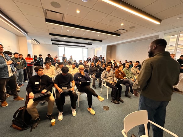 A man stands at the front of a modern indoor room, addressing a large group of people who are seated and standing, many of whom are using laptops.