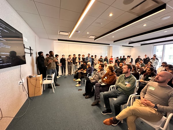 A man is giving a presentation to a large audience in a modern office setting, with code displayed on a large wall-mounted screen.