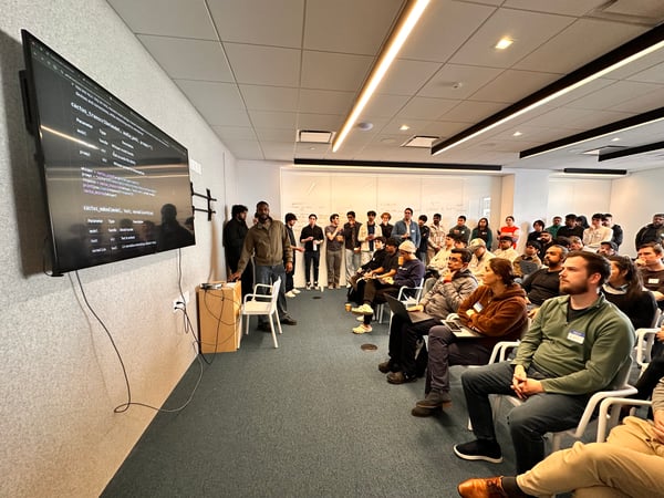 A man is giving a technical presentation to a large audience in a modern office space, with a screen displaying code examples.