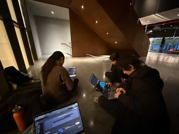 A group of three people are sitting on the floor of a modern building at night, focused on their open laptops.