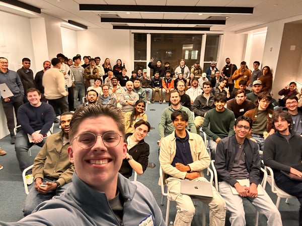 A large group of young adults, mostly men, are posing for a selfie in an indoor meeting room or classroom setting.
