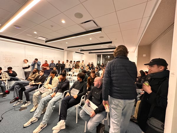 A group of people, mostly young men, are gathered in a modern office or classroom setting for a presentation or meeting. Some individuals are seated in rows of white chairs while others stand along the back and side walls.