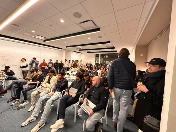 A group of people, mostly young men, are gathered in a modern office space for a presentation or meetup, with some seated in rows and others standing near a whiteboard.