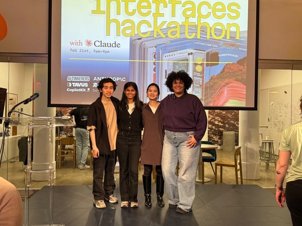 Four people pose for a photo on a stage in front of a projection screen advertising an 'Interfaces hackathon' with Claude.