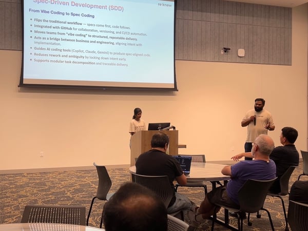 A photograph capturing a technical presentation indoors, showing two presenters (a woman at a podium and a man speaking to the audience) in front of a large screen displaying a slide about Spec-Driven Development (SDD). Several audience members are seated at round tables in the foreground.