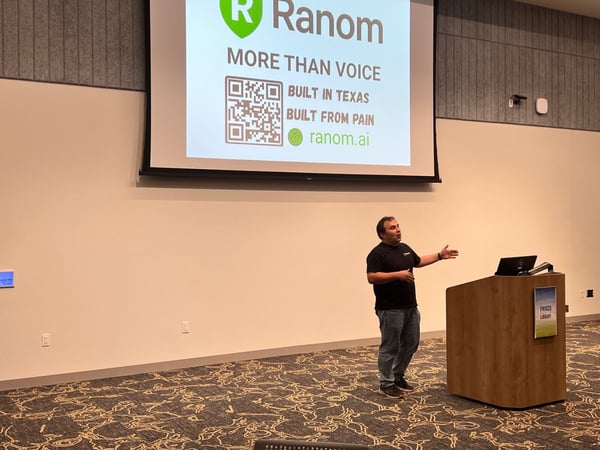 A man is giving a presentation in a conference room, standing near a wooden podium while gesturing toward a large screen displaying the 'Ranom' logo, a QR code, and promotional text.