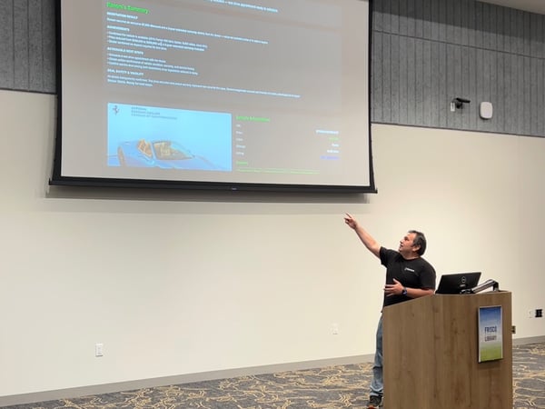 A man stands behind a wooden podium, giving a business presentation in a large indoor room and pointing toward a projection screen displaying detailed text about a vehicle negotiation result, including an image of a blue Ferrari.