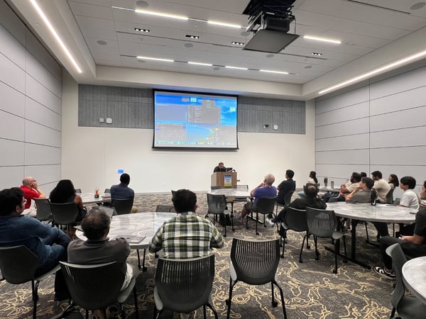 A photograph capturing a presentation or meeting in a large, modern conference room, showing a group of attendees seated at round tables facing a presenter and a large projection screen displaying technical content.
