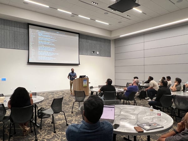 A photograph captures an indoor technical presentation where a speaker stands at a wooden podium next to a large projection screen displaying lines of code against a dark background, while an audience sits at round tables watching the event.