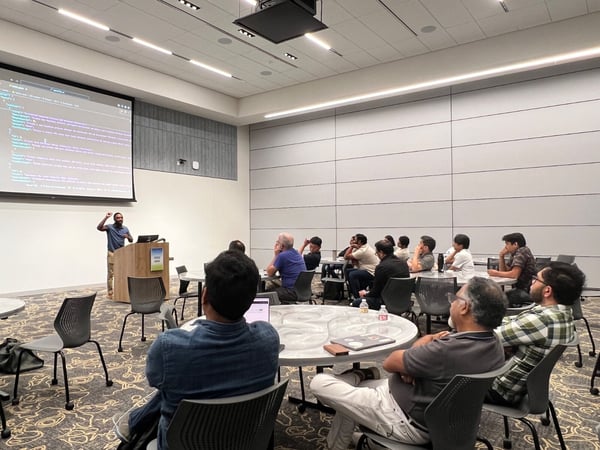 A photograph capturing a technical presentation in a modern conference room, showing a presenter standing at a podium and gesturing toward a large screen displaying code, while an audience is seated at round tables watching.