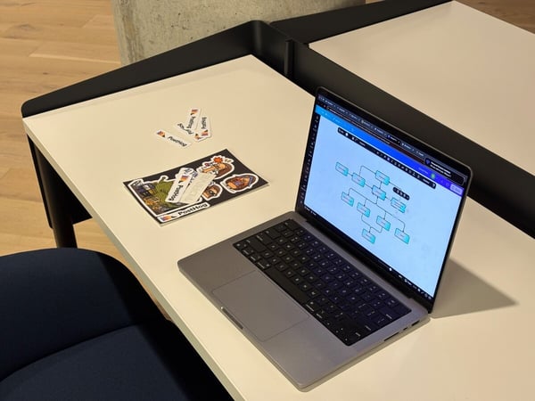 A close-up photograph of a modern workspace featuring an open laptop displaying a flowchart diagram, situated on a white desk next to a collection of PostHog branded stickers.
