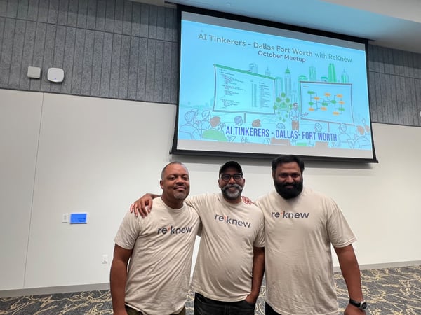 Three men wearing matching beige t-shirts branded with the text 're knew' pose together in an indoor event space in front of a projection screen displaying presentation slides for an AI Tinkerers meetup.