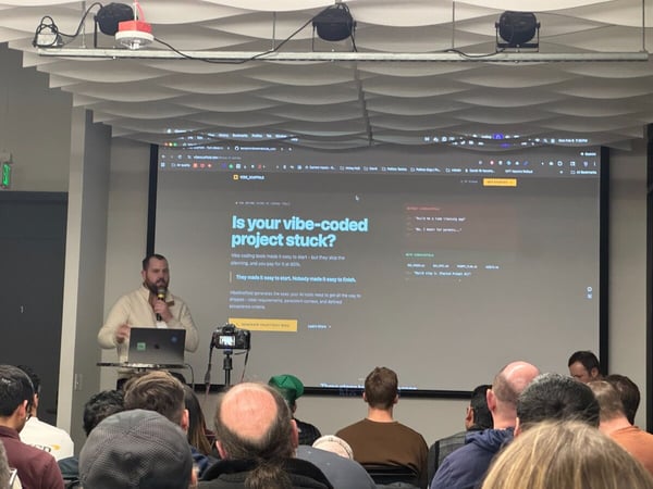 A man stands on a stage giving a presentation about 'Vibe Scaffold' to a seated audience, with a large screen displaying a website behind him.