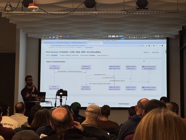 A man is giving a technical presentation to an audience, with a large screen displaying a diagram of 'Agent Communication' involving various AI models.