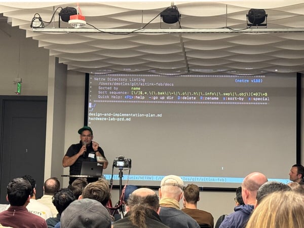 A man in a green cap is giving a presentation in front of a large screen displaying a terminal directory listing to an audience.