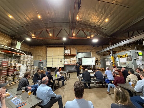 A group of people are gathered in a large brewery or warehouse space, sitting at picnic tables and watching a presentation on a large screen.