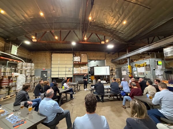 A group of people are seated at picnic tables in a large industrial brewery or warehouse, watching a presentation on a projector screen.