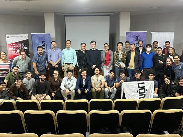 A group photo of approximately 33 people posing in an auditorium, featuring banners for a Master's in Artificial Intelligence program.