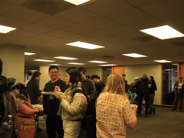 A group of people are gathered in an indoor office or event space, socializing and eating food during a gathering.
