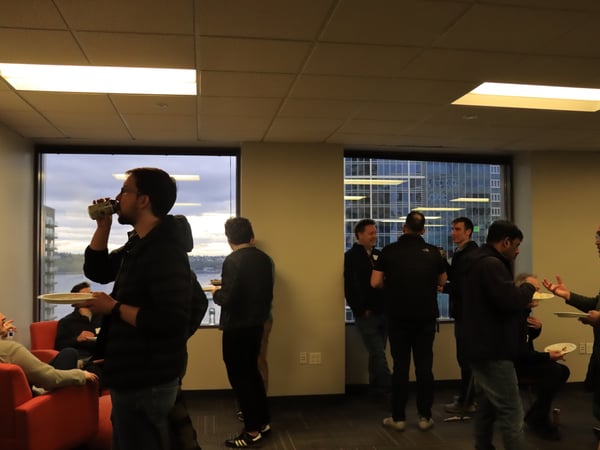 A group of men are socializing and networking in an office setting during the evening, with some eating and drinking while others engage in conversation.