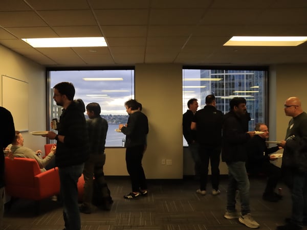 A group of people are gathered in an office space for a social event, standing near large windows overlooking a city and water at dusk.