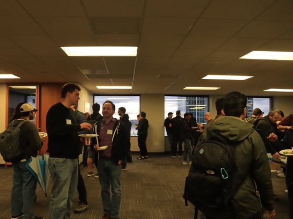 A group of people are gathered in a spacious office environment for a social or networking event, with several individuals holding plates of food.