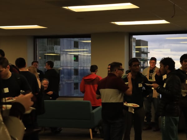 A group of people are gathered indoors for a social event, standing and eating from plates in a room with large windows overlooking a city.