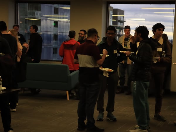 A group of people are gathered in an indoor office or event space, socializing and eating while standing near large windows.