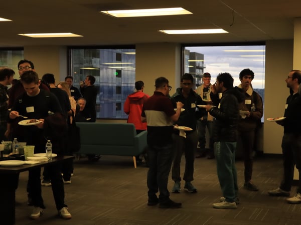 A group of people are gathered in an indoor office setting for a social event, eating food and talking near large windows overlooking a city at dusk.
