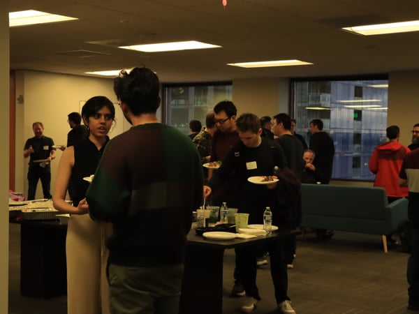 A group of people are gathered in an indoor office environment, socializing and eating food from a buffet table.