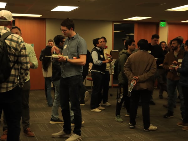 A group of people are gathered in an indoor office setting for a social event, with several individuals eating and others engaged in conversation.