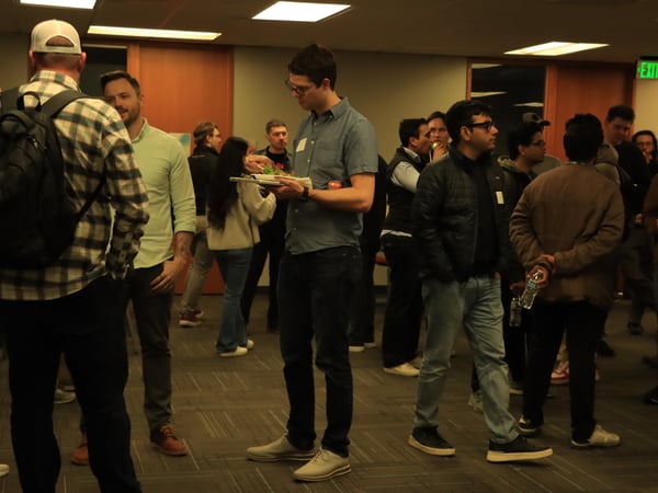 A group of people are gathered in an indoor setting for a social or networking event, with several individuals holding plates of food and drinks while conversing.