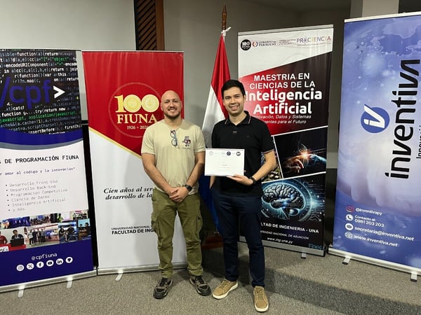 Two men stand in front of several promotional banners and a Paraguayan flag, with one man holding a certificate.