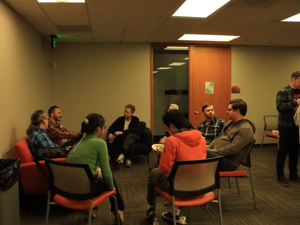 A group of people are gathered in a circle in an office or common room, sitting on a red couch and chairs while engaged in conversation.