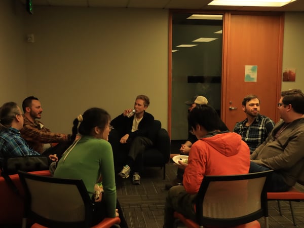 A group of people are sitting in a circle in an indoor office-like setting, engaged in a casual conversation while some are eating and drinking.