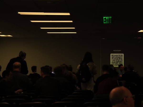 A dark auditorium filled with people attending a presentation, where a projector screen displays a QR code and the text 'Join the Survey!'.