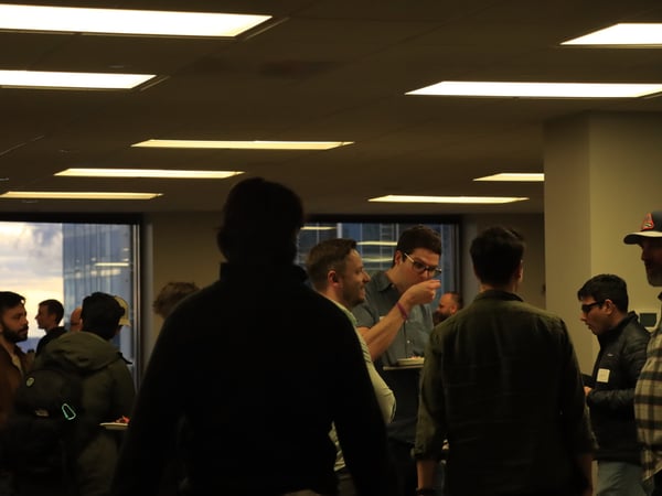 A group of people are gathered in an office-like setting for a social event, with some individuals eating and talking near a large window.