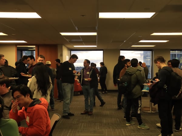 A candid photograph of a group of people networking and eating in a modern office space with large windows overlooking a city at dusk.