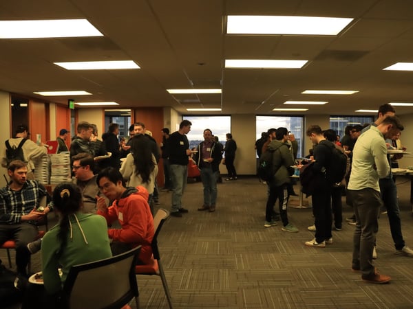 A group of people are gathered in a brightly lit office space for a social event, many of them holding plates of food and drinks.