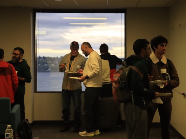 A group of men are gathered indoors for a social event, some holding plates of food, with a large window in the background showing a view of water and a cloudy sky.