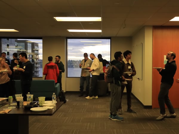 A group of people are gathered in an office setting for a social event, many of whom are holding plates of food and talking.