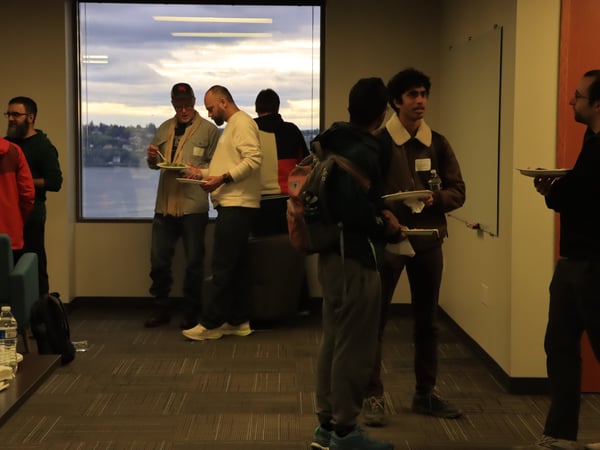 A group of men are socializing and eating in an office setting with a large window overlooking a body of water at dusk.