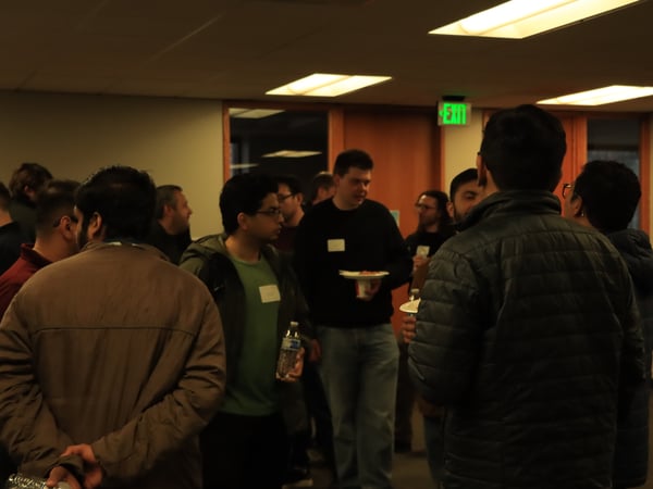 A group of people are gathered in an indoor office or event space, socializing in small groups with some individuals holding food and water.
