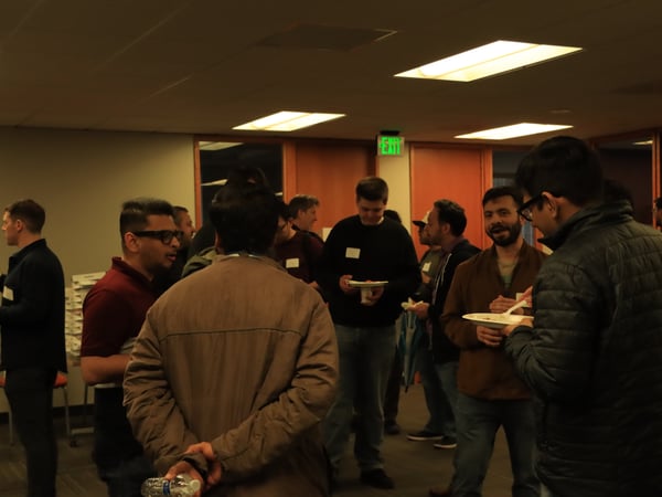 A group of men are gathered in a dimly lit indoor room, socializing and eating from paper plates during a networking event.