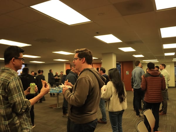 A group of people are gathered in an office-like setting, socializing while eating pizza and drinking from cans.