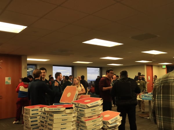 A group of people are gathered in an office setting for a social event, with several stacks of Domino's pizza boxes in the foreground.