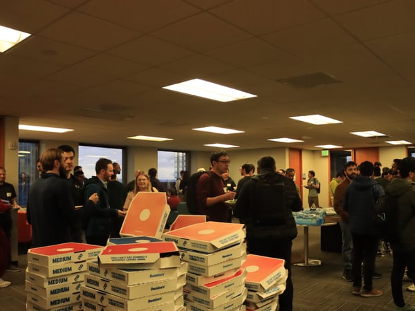A group of people gathering in an office-like setting for a social event with several stacks of Domino's pizza boxes in the foreground.