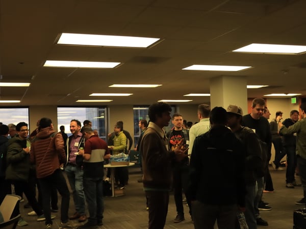 A group of people are gathered in a large office space for a social or networking event, standing in small groups and talking.