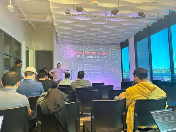 An indoor photograph of a tech meetup presentation, featuring a presenter standing near a projection screen displaying an agenda for 'May Demo Night Ft. Oracle & NVIDIA' to a seated audience.