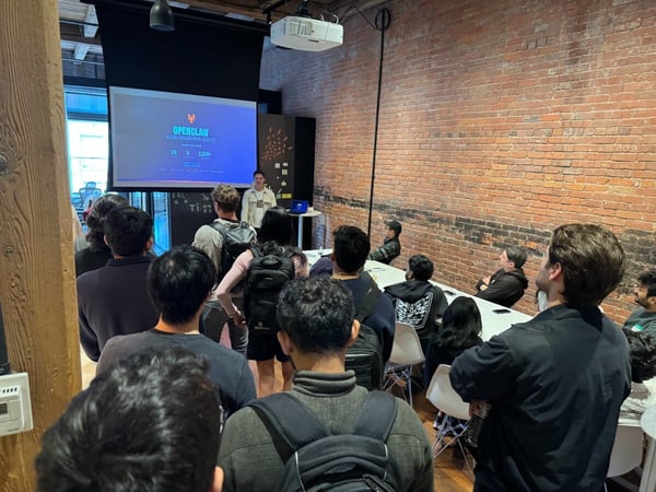 A group of people are gathered in a room with an exposed brick wall, watching a presentation on a screen titled 'OPENCLAW'. A speaker is standing at the front of the room near the projector screen.